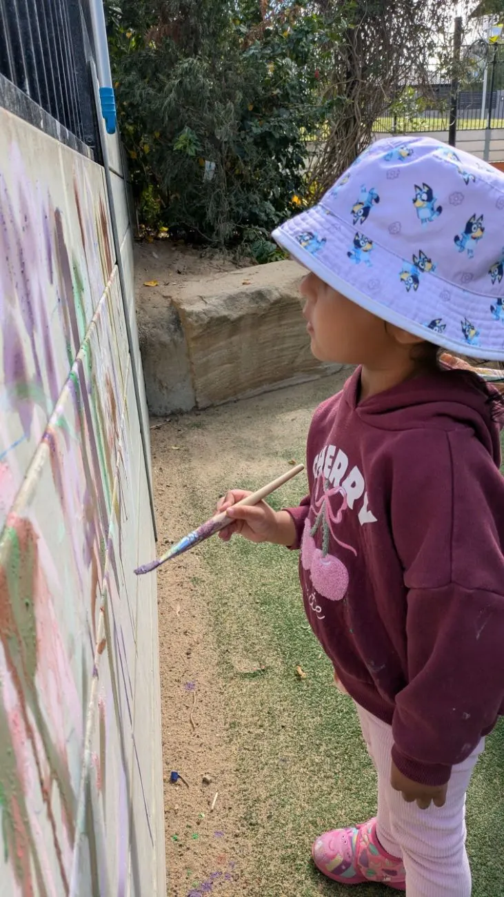 A child painting the outdoor fence canvas with a long brush