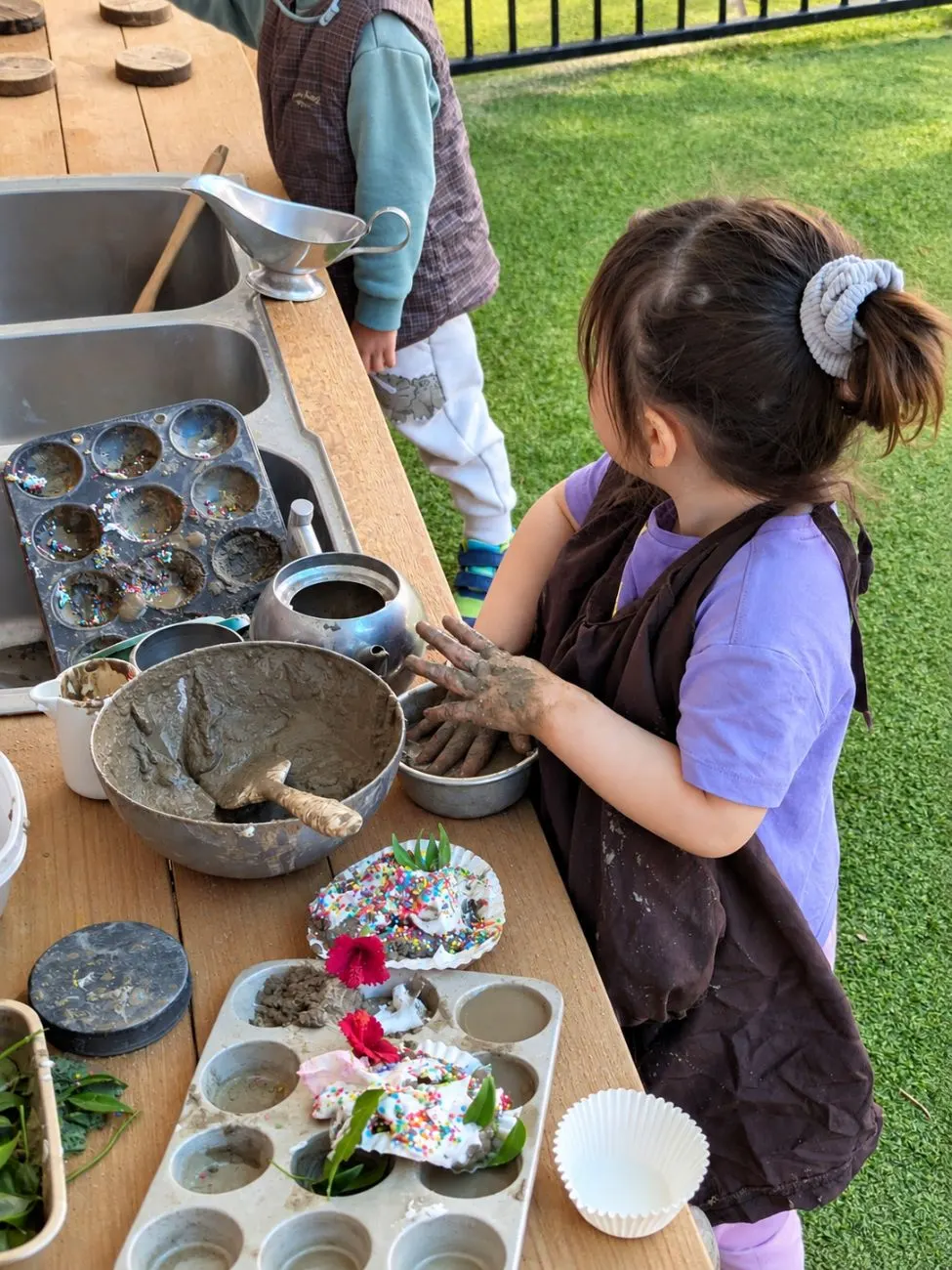A child in an apron making mud cakes with petals and sprinkles at the outdoor kitchen