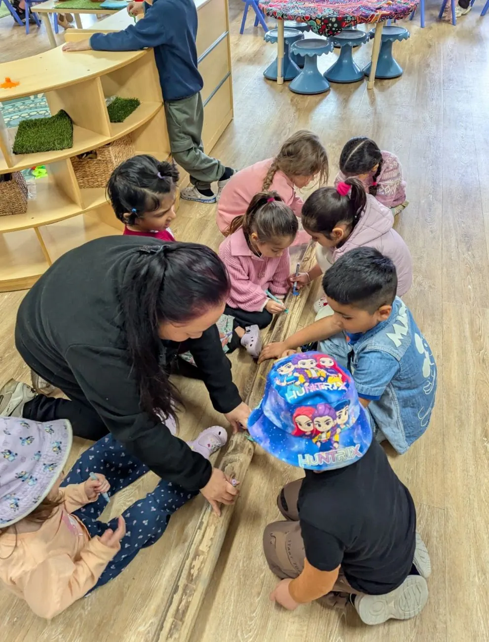 Children drawing along a timber log on the floor with an educator