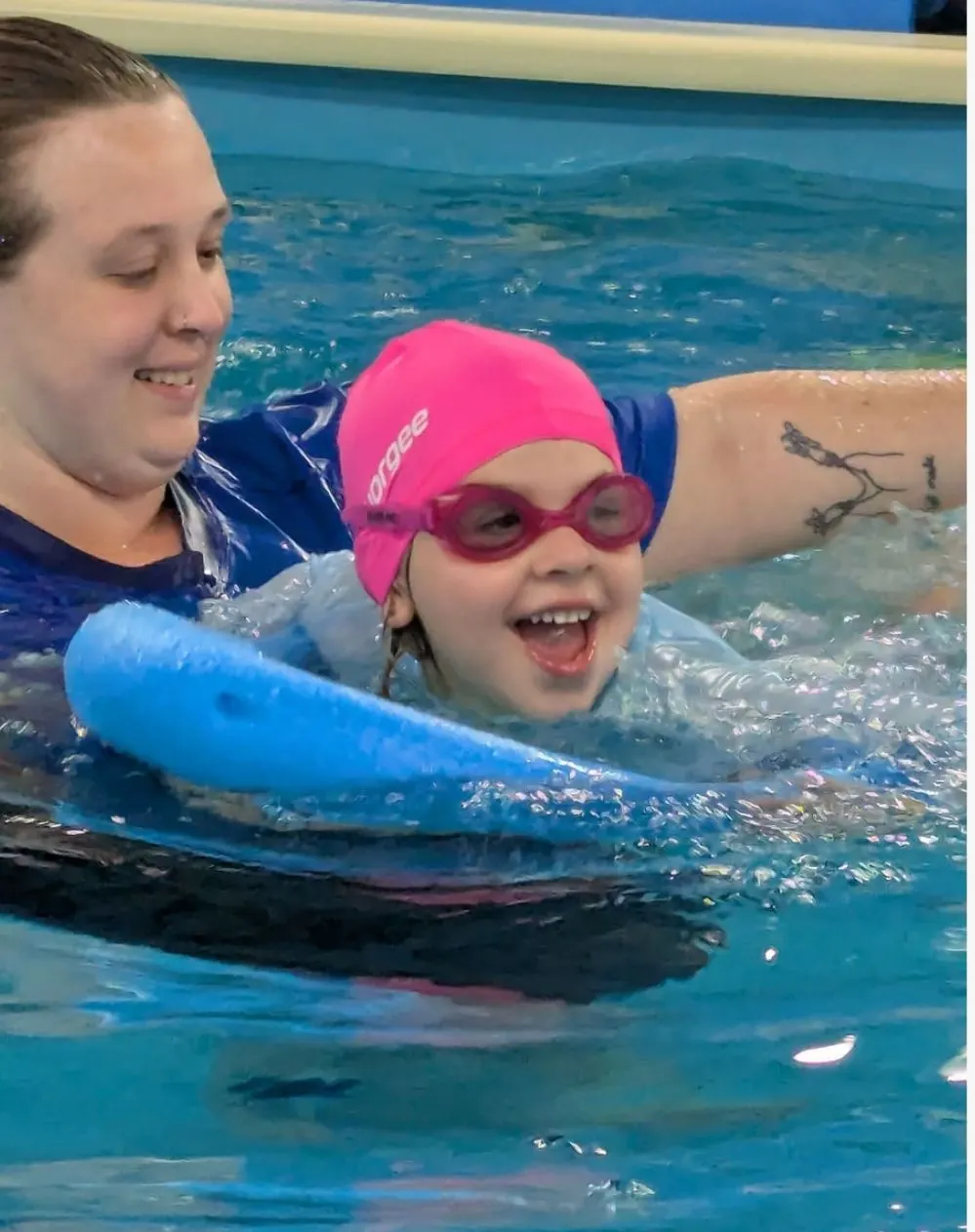 A child kicking along with a pool noodle and her swim teacher