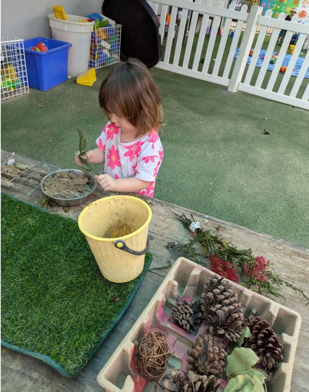 A toddler sorting leaves, pine cones and sand at the outdoor bench