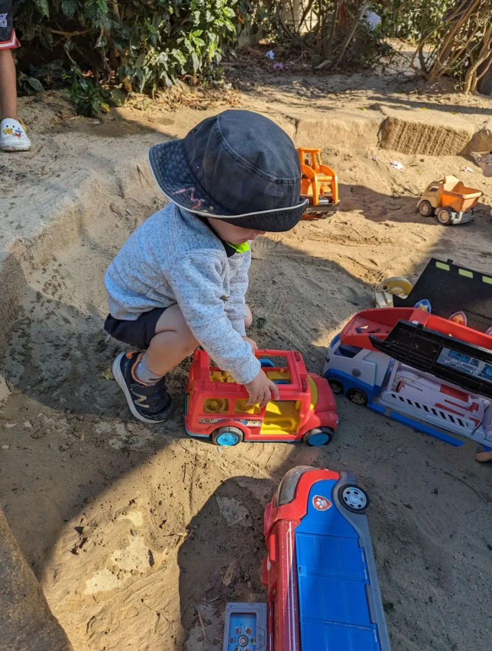 A toddler pushing trucks through the sandpit